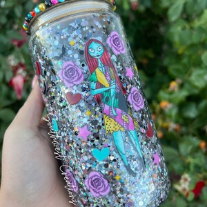 Puede incluir: Un vaso de purpurina plateado con una imagen en blanco y negro de Sally de Pesadilla Antes de Navidad. El vaso est&aacute; decorado con rosas moradas, corazones rojos, corazones turquesa y estrellas plateadas. El texto "Southwest Creations" est&aacute; en el lateral del vaso.