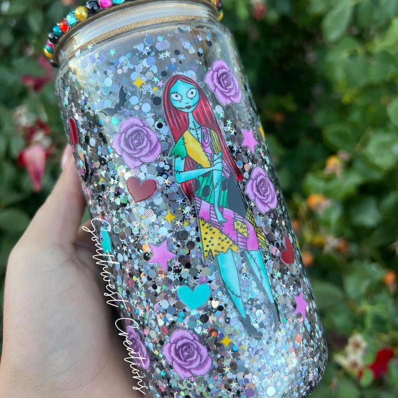 Puede incluir: Un vaso de purpurina plateado con una imagen en blanco y negro de Sally de Pesadilla Antes de Navidad. El vaso est&aacute; decorado con rosas moradas, corazones rojos, corazones turquesa y estrellas plateadas. El texto "Southwest Creations" est&aacute; en el lateral del vaso.