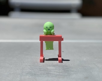 May include: A green figurine with a surprised expression is perched on top of a pink miniature hurdle.