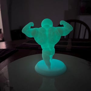 May include: A glowing, teal-colored figurine of a bodybuilder flexing his muscles. The figure is on a round, white base. The image is taken in a dimly lit room, with the figure's glow providing the primary light source.