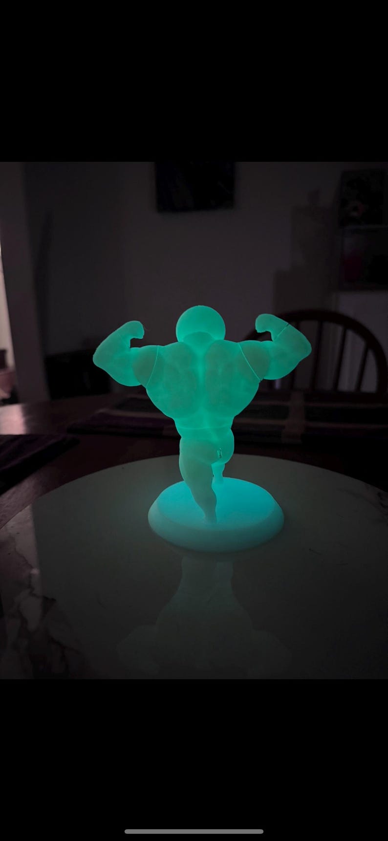 May include: A glowing, teal-colored figurine of a bodybuilder flexing his muscles. The figure is on a round, white base. The image is taken in a dimly lit room, with the figure's glow providing the primary light source.