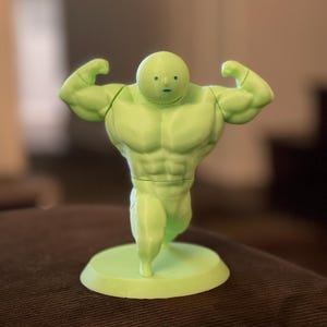 May include: A light green figurine of a muscular figure with a small head and arms flexed. The figurine is standing on a circular base. The figure has a cartoonish appearance and is made of a solid material.