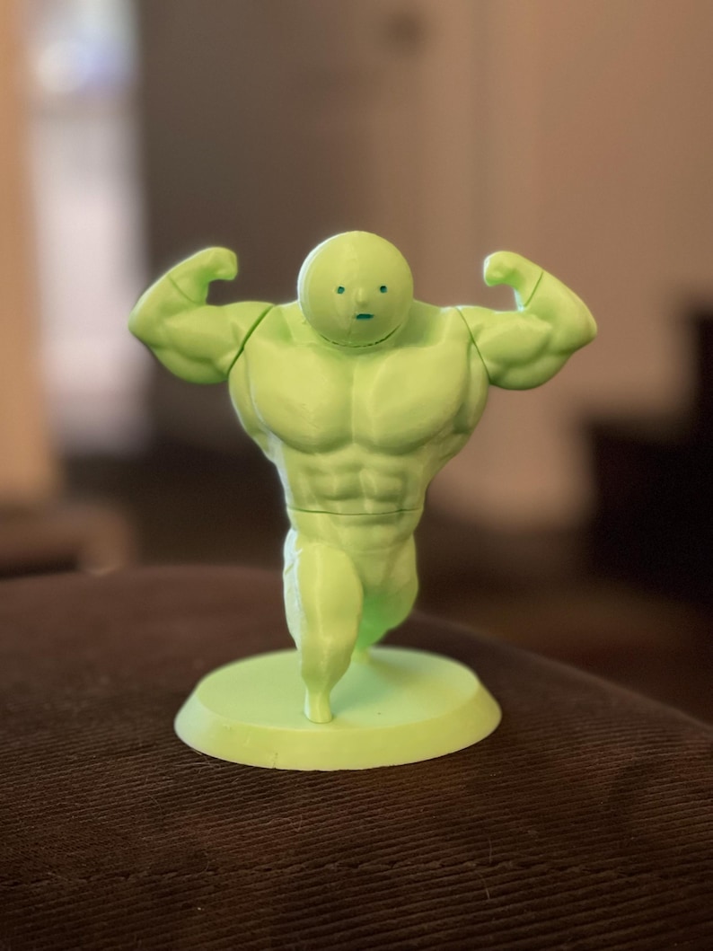 May include: A light green figurine of a muscular figure with a small head and arms flexed. The figurine is standing on a circular base. The figure has a cartoonish appearance and is made of a solid material.