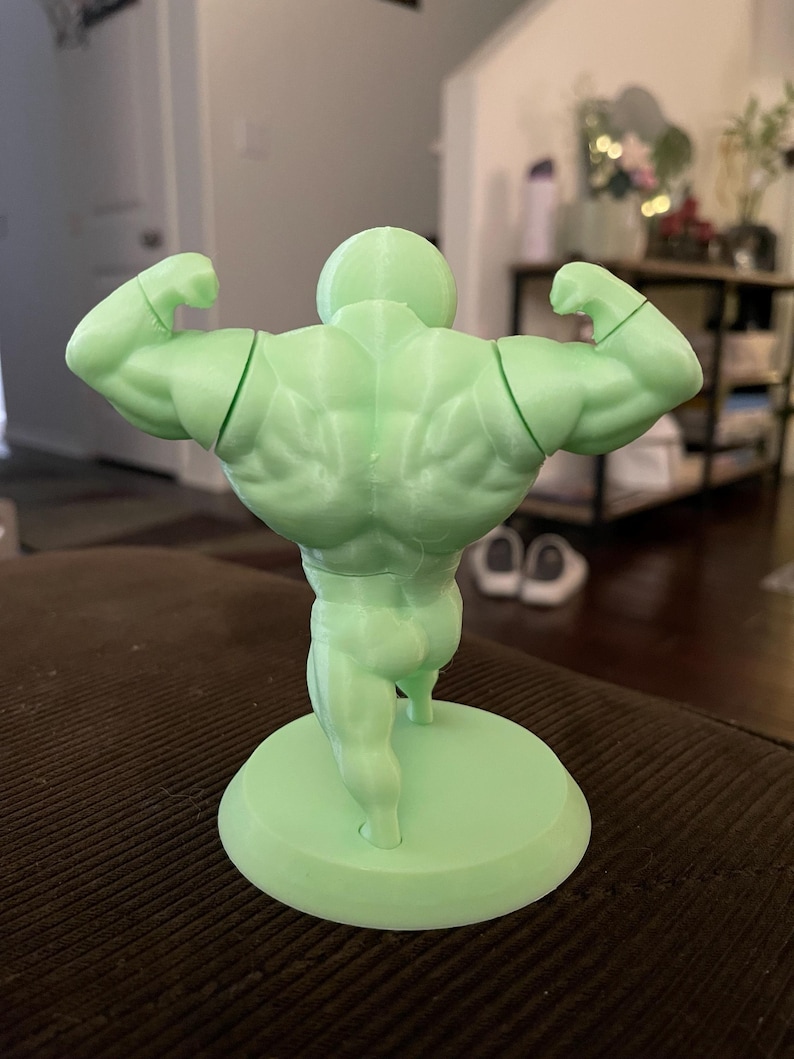 May include: A light green figurine of a bodybuilder flexing his muscles. The figurine is standing on a circular base. The figure's back muscles are clearly defined, and the arms are flexed in a classic pose.