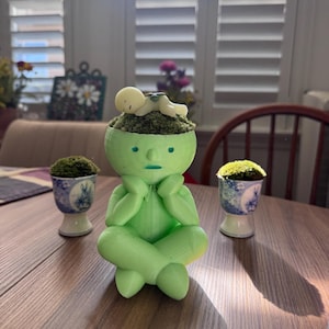 May include: A light green figurine of a person in a seated pose with a small figure lying on its head, surrounded by two small decorative pots with moss. The figurine has a serene expression.