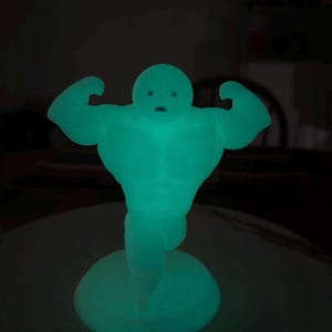 May include: A glowing teal figurine of a muscular figure flexing its arms. The figure has a round head with simple facial features and stands on a circular base. The object is illuminated in a dark setting.