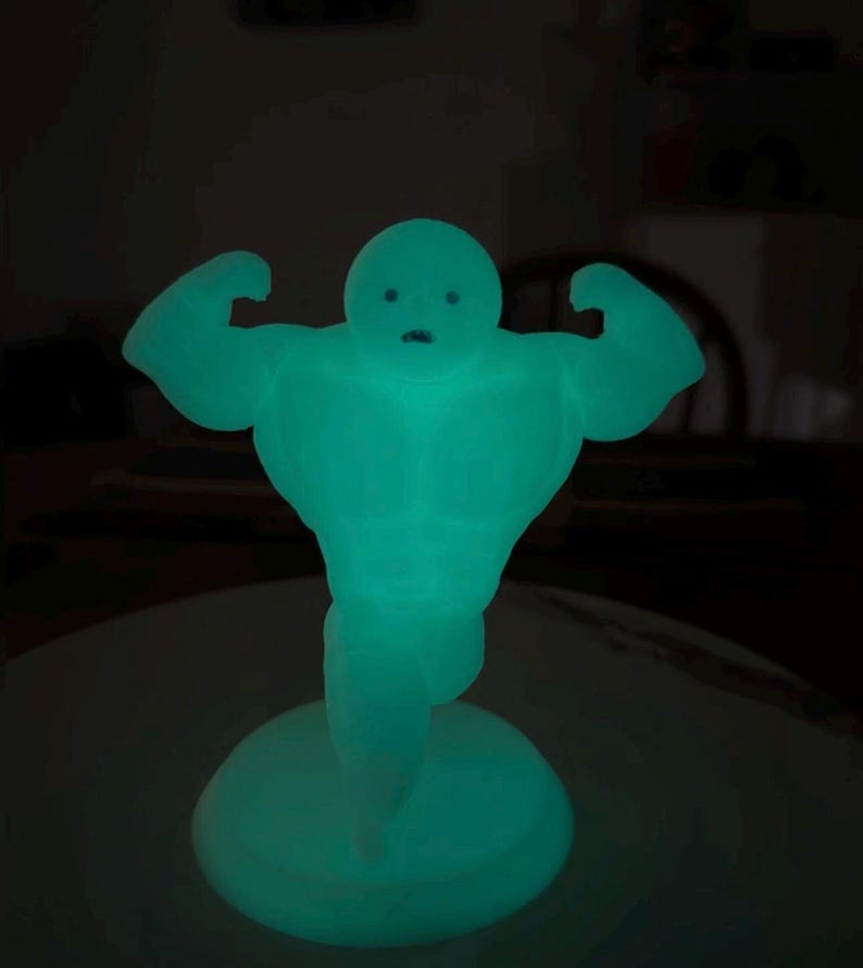 May include: A glowing teal figurine of a muscular figure flexing its arms. The figure has a round head with simple facial features and stands on a circular base. The object is illuminated in a dark setting.
