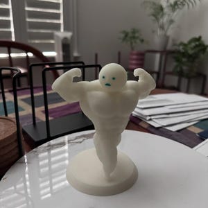 May include: A white figurine of a muscular figure flexing its biceps. The figure has a round head with small blue eyes and a small base. The figurine is on a white surface.