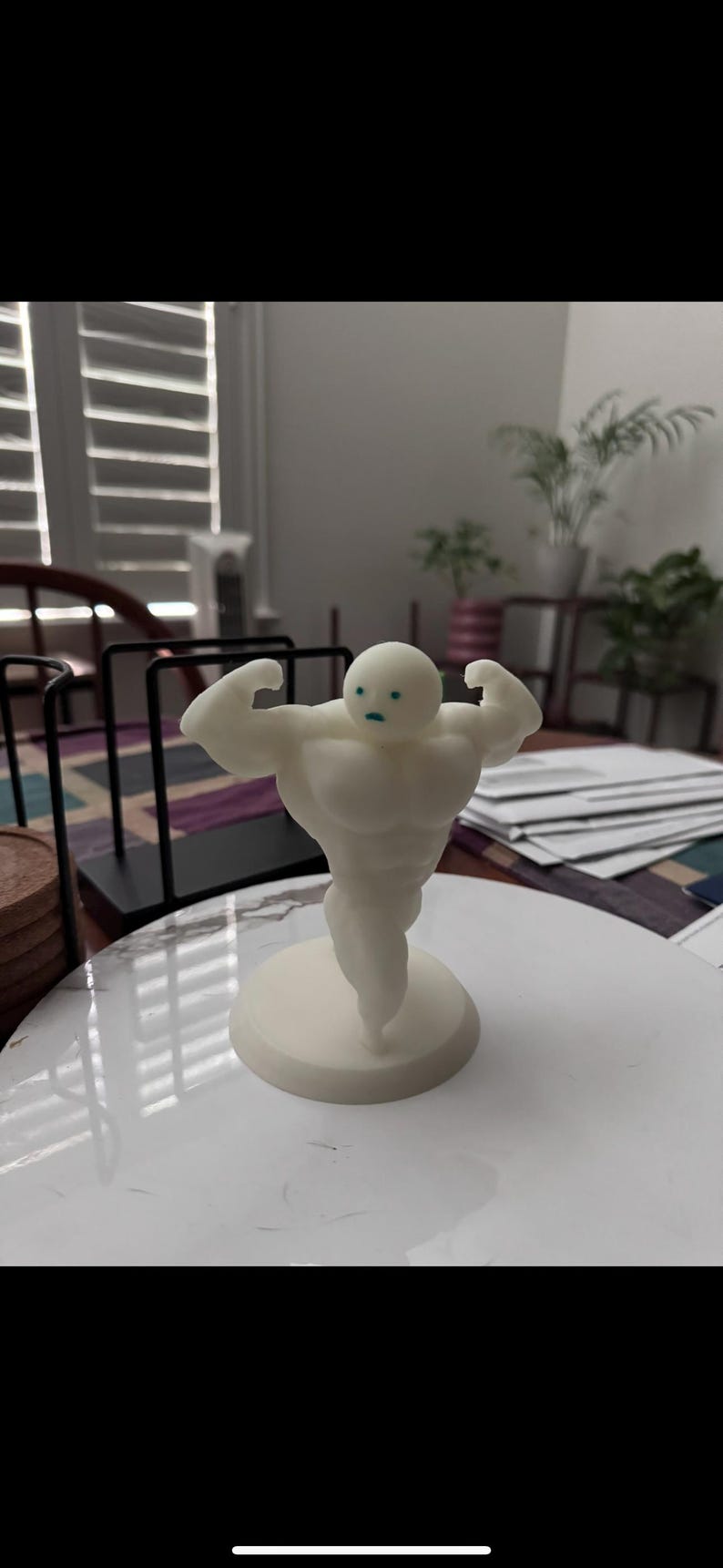 May include: A white figurine of a muscular figure flexing its biceps. The figure has a round head with small blue eyes and a small base. The figurine is on a white surface.