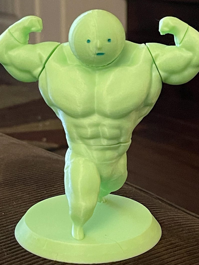 May include: A green 3D printed figurine of a muscular figure with a large head and small limbs. The figure is standing with its arms raised in a flexing pose.