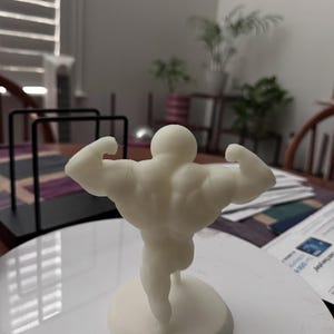 May include: A cream-colored figurine of a bodybuilder flexing his muscles. The figure is standing on a circular base. The background is blurred, but appears to be indoors.