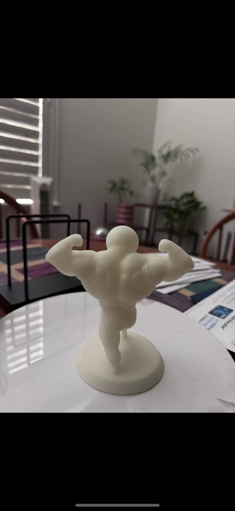 May include: A cream-colored figurine of a bodybuilder flexing his muscles. The figure is standing on a circular base. The background is blurred, but appears to be indoors.