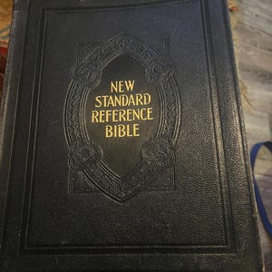 Könnte beinhalten: Ein schwarzes Hardcover-Buch mit dem Titel "New Standard Reference Bible" in goldener Schrift. Der Einband hat ein geprägtes Design um den Titel. Das Buch scheint abgenutzt zu sein, was darauf hindeutet, dass es sich um ein antikes oder Vintage-Exemplar handelt.