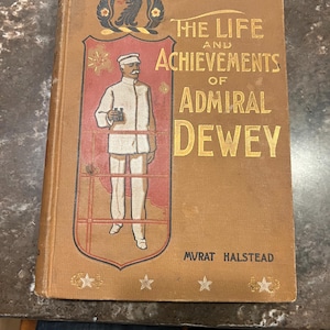 May include: Vintage book titled "The Life and Achievements of Admiral Dewey" by Murat Halstead. The cover is tan with gold lettering and a red and black illustration of a man in a white uniform. The book has decorative stars.