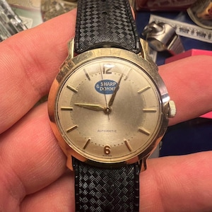 Puede incluir: Un reloj de pulsera automático Sharp & Dohme en tono dorado con una correa negra texturizada. La esfera del reloj es plateada con agujas e indicadores dorados. La esfera del reloj tiene el texto "SHARP & DOHME" y "AUTOMATIC".