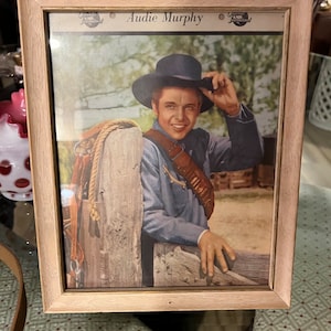 Puede incluir: Una fotografía enmarcada de Audie Murphy, un actor y soldado estadounidense, con un sombrero de vaquero y una camisa azul. La foto está en un marco de madera clara con un paspartú blanco.