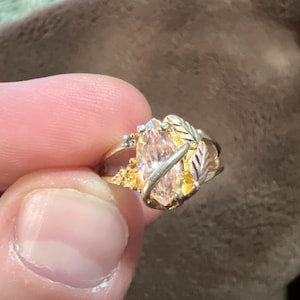 Op de afbeelding: Een gouden ring met een ovale, lichtroze edelsteen. De ring heeft een bladerontwerp aan de zijkanten en kleine accentstenen. De ring wordt tussen de vingers gehouden.