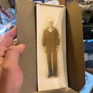 Puede incluir: Una fotografía antigua en blanco y negro de un marinero con uniforme, que incluye una gorra blanca y un jersey oscuro con cuello en pico. El retrato se presenta dentro de una carpeta de cartón gris texturizado que se abre para mostrar la imagen.