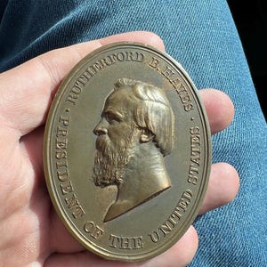 Può includere: Una medaglia di bronzo con un ritratto di profilo di Rutherford B. Hayes, il 19° presidente degli Stati Uniti. La medaglia è di forma ovale e ha l'iscrizione "Rutherford B. Hayes, Presidente degli Stati Uniti" attorno al bordo.