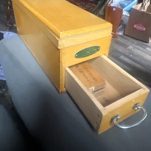 May include: A rectangular wooden box with a sliding drawer. The box is painted a light yellow color and has a metal handle on the drawer. A small green oval-shaped label is on the front of the box. The drawer is open, revealing a wooden block with Japanese text.