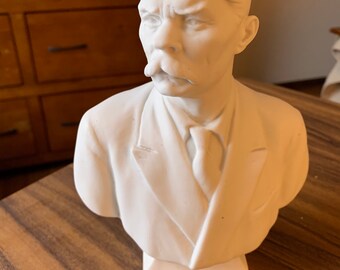 May include: White porcelain bust of a man with a mustache. The bust is on a white base.