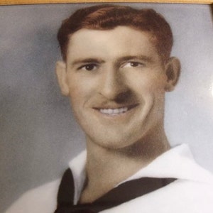 May include: A black and white photo of a man in a sailor uniform, smiling. He is wearing a white shirt with a black collar and a black tie. The photo is framed in a wooden frame.