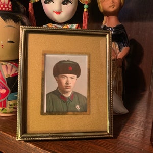 May include: A framed black and white photograph of a young man wearing a green military uniform and a black fur hat with a red star. The photo is displayed on a wooden shelf with two dolls.