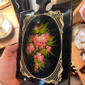 May include: Black wooden serving board with a handle, featuring a hand-painted floral design. The oval centerpiece showcases pink flowers and green leaves, framed by a gold filigree border. The board is rectangular, with a glossy finish.