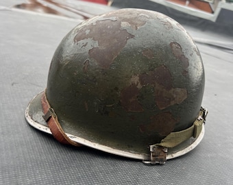 Könnte beinhalten: Ein olivgrüner Militärhelm mit einem braunen Lederkinnriemen und grünen Canvas-Riemen. Die Oberfläche des Helms weist Gebrauchsspuren auf, mit Bereichen, in denen die Farbe abgeblättert ist und das darunterliegende Metall freilegt. Ein Vintage-Militärsammlerstück.