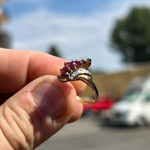 May include: A gold ring featuring a cluster of marquise-cut deep pink gemstones arranged in a fan-like pattern, accented by a small row of clear stones set into the band, showcasing a polished metal finish and a curved, bypass-style shank design.