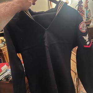 Peut inclure: Chemise de marin vintage bleu marine avec un large col et des rayures blanches. La chemise présente des écussons sur la manche avec un aigle et d'autres insignes. La chemise est suspendue à un cintre en bois.