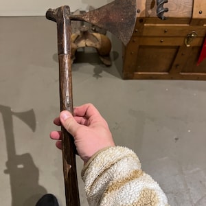 Puede incluir: Un hacha antigua con un mango de madera oscura y una hoja de metal oxidado. La cabeza del hacha está unida al mango con una cuña de madera. El mango está tallado con un patrón texturizado. El hacha está en la mano de una persona.
