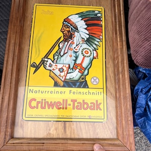 May include: Framed vintage advertisement featuring a Native American in a headdress, holding a pipe. The yellow background displays the text "Jnka" and "Crüwell-Tabak" in red and black. The wooden frame adds a classic touch to this piece.