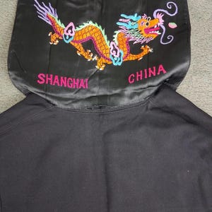 May include: Black garment with an embroidered dragon design in orange, yellow, pink, and blue. The text "SHANGHAI CHINA" is embroidered in pink. The garment appears to be a hood or collar.