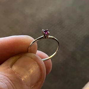 May include: A thin gold ring featuring a small, round, deep pink gemstone set in a four-prong setting. The band is smooth and slender, showcasing a minimalist design that highlights the vibrant color of the central stone held in the metal prongs.