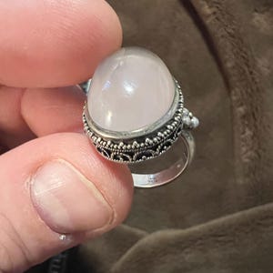May include: A silver ring featuring a large, light pink, rounded gemstone. The ring has an ornate, filigree design around the stone and a simple band. The gemstone is smooth and polished.