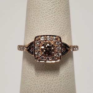 Puede incluir: Anillo de oro rosa con una piedra central cuadrada rodeada de piedras blancas más pequeñas. El anillo tiene detalles negros a ambos lados de la piedra central y una banda con pequeñas piedras blancas.