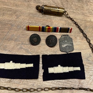 May include: A collection of military memorabilia including a chain, a medal, two buttons, a ribbon, and two fabric patches with embroidered ship silhouettes. The medal is silver and features the text "The Naval First National Bank of America".