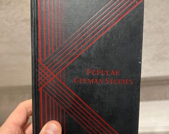 Pode incluir: Um livro vintage intitulado "Popular German Stories". A capa é preta com um design geométrico vermelho e texto vermelho. O livro é segurado em uma mão.