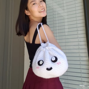 May include: A white crocheted bag shaped like a steamed bun with a smiling face and pink cheeks. The bag has black eyes and a black mouth.