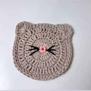 May include: A beige crocheted cat face with a pink nose and black whiskers. The crocheted cat face is on a white background.