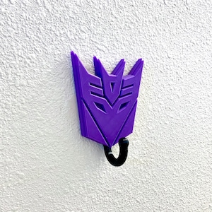 May include: Purple plastic wall hook shaped like the Transformers character Megatron's symbol. The hook has a black metal hook at the bottom.