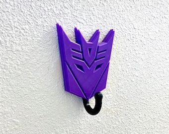 May include: Purple plastic wall hook shaped like the Transformers character Megatron's symbol. The hook has a black metal hook at the bottom.