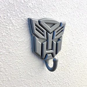 May include: A gray 3D printed wall hook shaped like the Transformers logo. The hook has a single hook at the bottom.