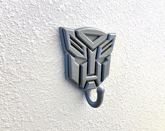 May include: A gray 3D printed wall hook shaped like the Transformers logo. The hook has a single hook at the bottom.