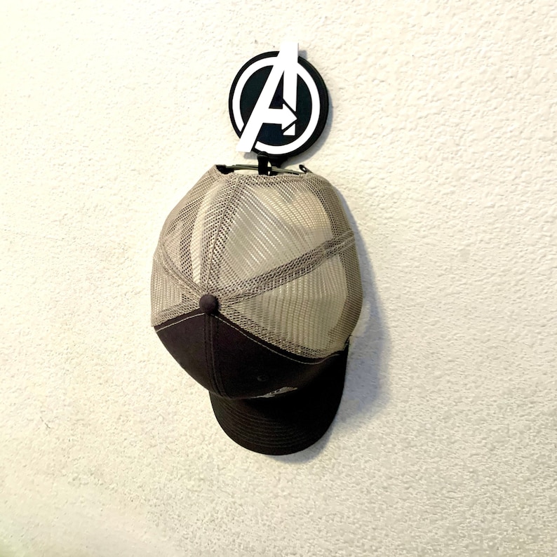 May include: A white plastic wall hook shaped like the Avengers "A" logo with a brown and beige baseball cap hanging from it.