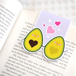 May include: A purple bookmark with two avocado halves with smiling faces and hearts. The avocado halves are holding hands. There are pink hearts floating around the avocados.