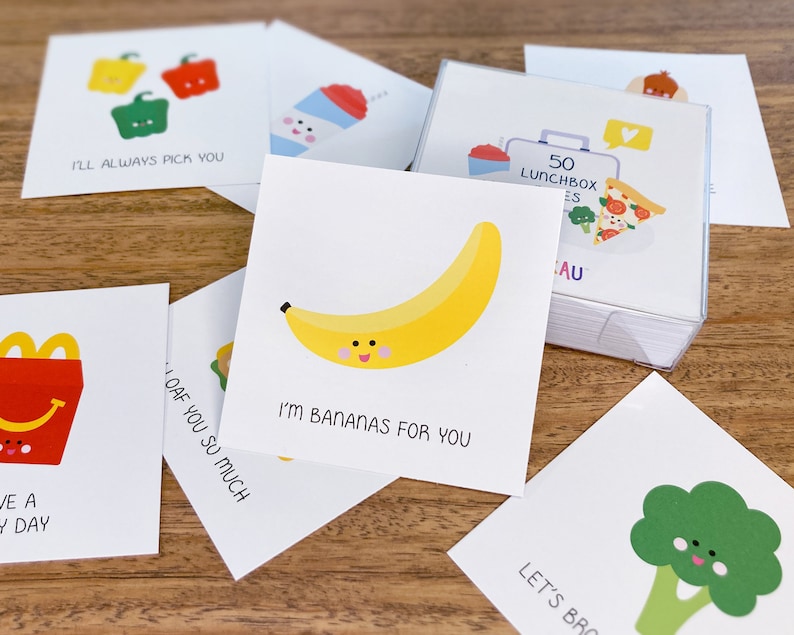 May include: A set of 50 cards with cute food illustrations and puns. The cards feature a banana, a broccoli, a pepper, a french fry, and a loaf of bread. The text on the cards includes phrases like "I'm bananas for you" and "Let's broc-oli!"