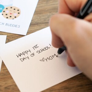 May include: A white card with the text "Happy 1st Day of School!" written in black ink. The card also has a heart symbol and the word "MOM" written in black ink.