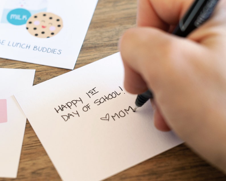 May include: A white card with the text "Happy 1st Day of School!" written in black ink. The card also has a heart symbol and the word "MOM" written in black ink.